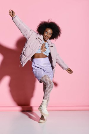A stylish young African American woman with curly hair joyfully dances in a vibrant pink room.