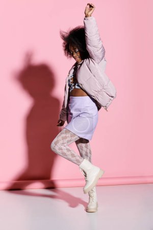 A young African American woman with curly hair energetically dancing in front of a vibrant pink wall in a stylish, modern studio.