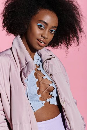A stylish young African American woman with curly hair wearing a pink jacket and a blue shirt in a studio setting.