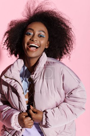 A stylish young African American woman with curly hair wearing a jacket and a blue makeup poses confidently in a studio.