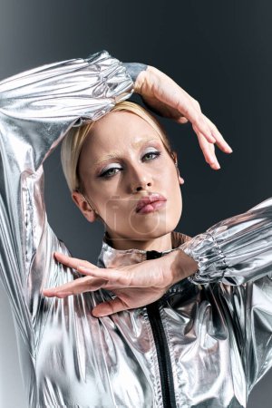 Téléchargez les photos : Séduisante femme aux cheveux blonds en tenue futuriste argentée posant en mouvement sur fond gris - en image libre de droit