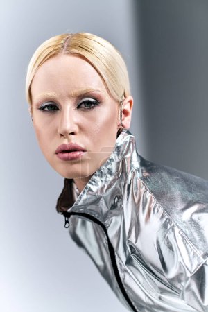 Téléchargez les photos : Séduisante femme aux cheveux blonds en tenue futuriste argentée posant en mouvement sur fond gris - en image libre de droit
