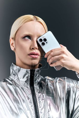 atractiva mujer en traje de plata futurista posando con el teléfono y mirando hacia otro lado en el fondo gris