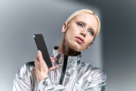 attraktive Frau im futuristischen silbernen Outfit posiert mit Handy und schaut vor grauem Hintergrund weg