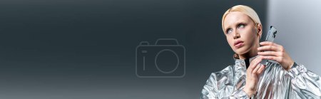 Téléchargez les photos : Belle femme en tenue futuriste posant avec téléphone et regardant loin sur fond gris, bannière - en image libre de droit