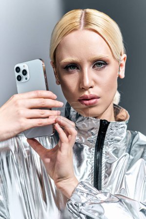 Téléchargez les photos : Séduisante femme extravagante en tenue futuriste argentée posant avec téléphone et regardant la caméra - en image libre de droit