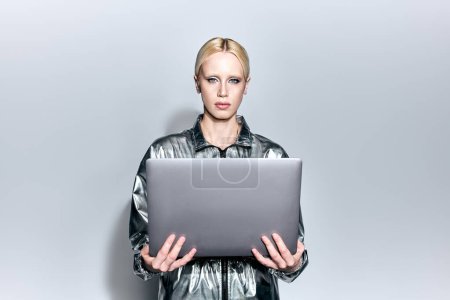 Téléchargez les photos : Belle blonde modèle féminin en tenue robotique argent tenant ordinateur portable et regardant la caméra - en image libre de droit
