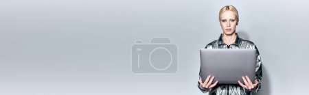 Photo for Beautiful blonde female model in robotic silver outfit holding laptop and looking at camera, banner - Royalty Free Image