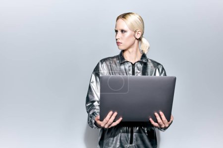 Téléchargez les photos : Extravagante belle femme en argent vêtements robotiques posant en mouvement avec ordinateur portable et détournant les yeux - en image libre de droit
