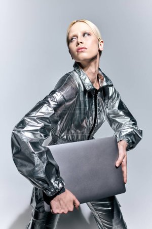 mujer atractiva extravagante en la ropa robótica de plata posando en movimiento con el ordenador portátil y mirando hacia otro lado