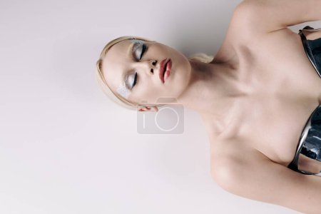 Téléchargez les photos : Séduisante femme bizarre aux cheveux blonds et maquillage en tenue futuriste argentée couchée sur le sol - en image libre de droit
