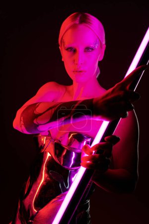 mujer peculiar en traje futurista metálico sosteniendo palo de lámpara led rosa y mirando a la cámara