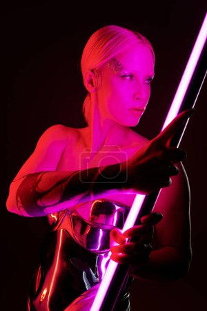 atractiva mujer en traje futurista metálico sosteniendo palo de lámpara led rosa y mirando hacia otro lado