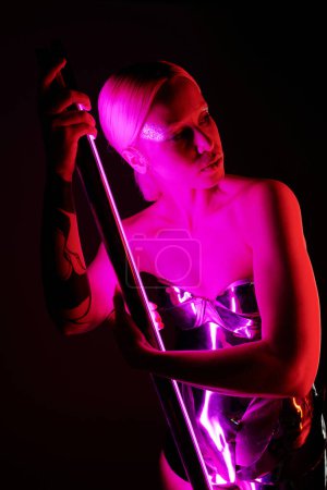 attraktive sonderbare Frau in futuristischer metallischer Kleidung, die einen rosafarbenen LED-Lampenstock hält und wegschaut