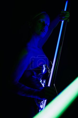 atractiva mujer extraña en traje robótico posando con palo de lámpara led azul y mirando a la cámara