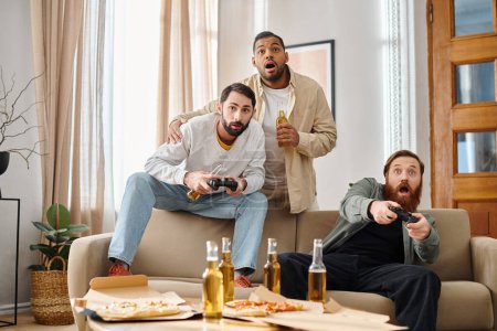Three cheerful, handsome men of different ethnicities enjoy a gaming session on a couch, displaying camaraderie and joy.