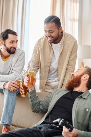 Tres hombres alegres y guapos de diferentes razas en atuendo casual, uniéndose y pasándola muy bien sentados encima de un sofá.