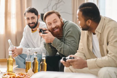 Téléchargez les photos : Trois beaux hommes de races différentes s'assoient autour d'une table, riant et se réjouissant mutuellement en compagnie pendant qu'ils tiennent des joysyicks. - en image libre de droit