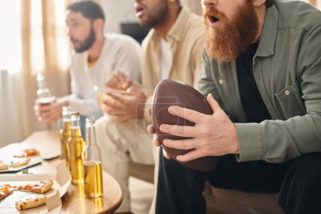 Three cheerful, interracial men in casual attire sit around a table, engrossed in a football game.