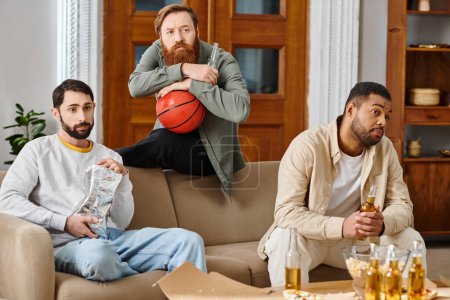Three interracial, handsome men sitting on a couch, enjoying drinks and watching basketball together in casual attire, showcasing friendship.