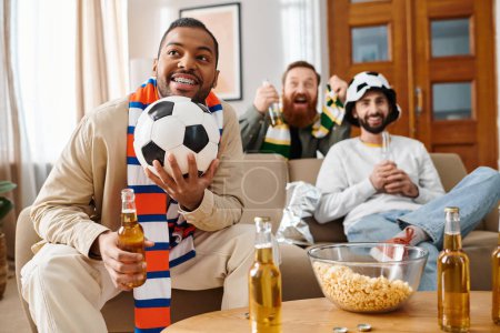 Foto de Un hombre con atuendo casual sostiene una botella de cerveza mientras sostiene una pelota de fútbol, disfrutando de un momento alegre con amigos en casa. - Imagen libre de derechos
