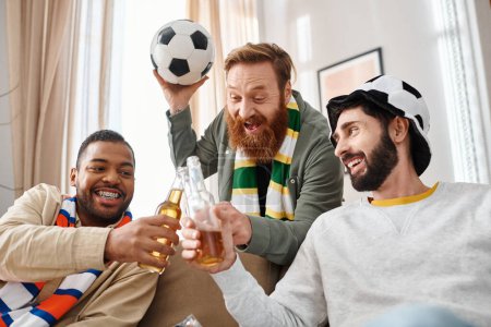 Three cheerful, interracial men in casual attire, bonding over a soccer ball on a cozy couch at home.