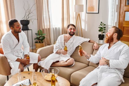 Three diverse, cheerful men wearing bathrobes have a great time chatting and laughing while sitting on top of a couch.