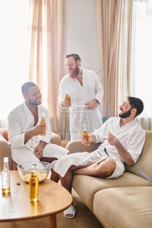 Three diverse, cheerful men in bathrobes enjoy each others company while seated on a couch.