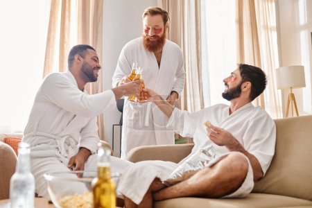Foto de Tres hombres diversos en albornoces riendo y charlando en un acogedor ambiente de sala de estar. - Imagen libre de derechos