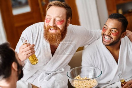 Diverse group of three cheerful men in bathrobes sitting at table, laughing and sharing a bowl of popcorn.