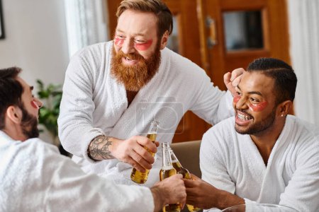 Foto de Tres hombres alegres en albornoces disfrutando de bebidas y camaradería alrededor de una mesa. - Imagen libre de derechos
