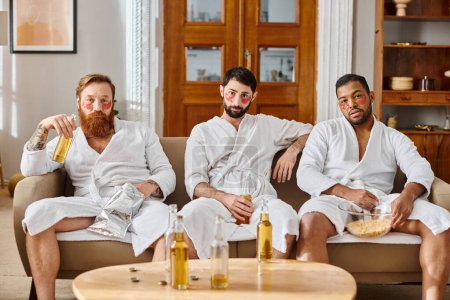 Three diverse, cheerful men wearing bathrobes, sitting on top of a couch, enjoying a great time together.