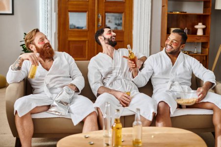 Three diverse men, wearing bathrobes, sit on top of a couch, smiling and chatting together in a cozy setting.