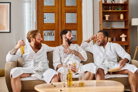 Three diverse men in bathrobes sit on top of a couch, chatting and laughing, forming a strong bond of friendship.