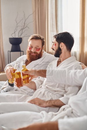 Three diverse and cheerful men in bathrobes enjoy each others company as they sit on top of a sofa, exuding a sense of camaraderie.