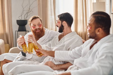 Foto de Tres hombres alegres en albornoces se sientan en un sofá, compartiendo risas y bebidas mientras disfrutan de la compañía de los demás. - Imagen libre de derechos