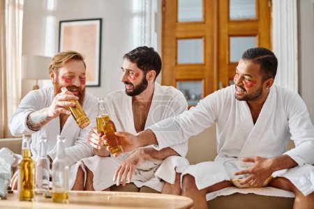 Diverse men in bathrobes sit on a couch, laughing and enjoying drinks from bottles in a cozy setting.