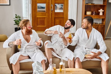 Three diverse men in bathrobes sit on the sofa, laughing and enjoying each others company in a joyful moment.
