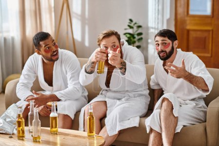 Three cheerful, diverse men in bathrobes sit on top of a couch, sharing laughter and camaraderie.