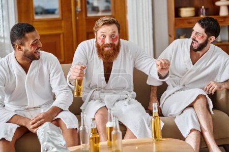 Three diverse cheerful men in bathrobes having a great time lounging and laughing on top of a couch.