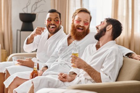 Three cheerful, diverse men in bathrobes share laughter and camaraderie while sitting on top of a plush couch.