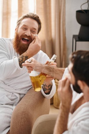 A man in a bathrobe sits on a couch, holding a glass of beer, deep in contemplation next to his friend.