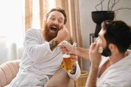 A man, wearing a bathrobe, relaxes while joyfully holding a beer next to his friend in a cheerful gathering.