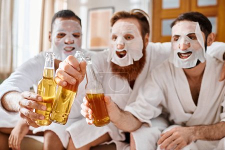 Three diverse, cheerful men in bathrobes, wearing facial masks, enjoy a fun moment together, clinking glasses of beer.