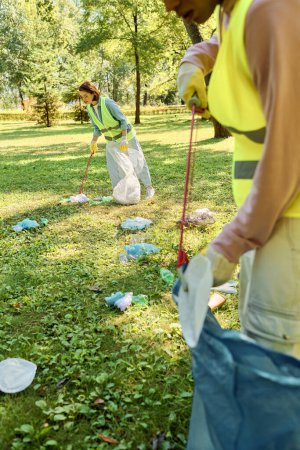 A socially active, diverse loving couple in safety vests and gloves clean a park as a group stands on a lush green field.