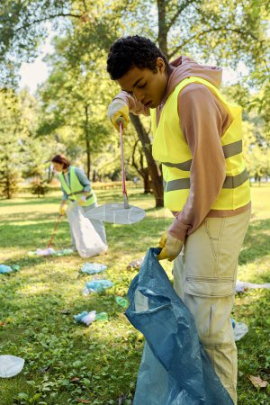 Diversa pareja en un chaleco amarillo está sosteniendo una bolsa azul mientras participa en una limpieza del parque con una pareja amorosa diversa socialmente activa.