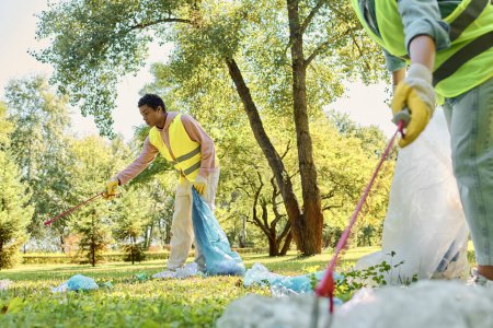 Una pareja diversa, con chalecos y guantes de seguridad, trabajando juntos para limpiar un parque cubierto de hierba.
