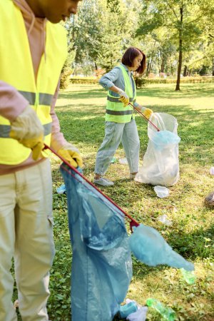 Couple socialement actif dans des gilets de sécurité et des gants travaillant ensemble pour nettoyer le parc, tenant des sacs poubelle.