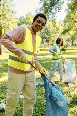 Diverse couple in a yellow vests holding a blue bag while cleaning a park