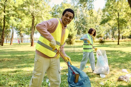 Hombre afroamericano en un chaleco de seguridad amarillo sostiene una bolsa azul mientras limpia en un parque con su esposa
